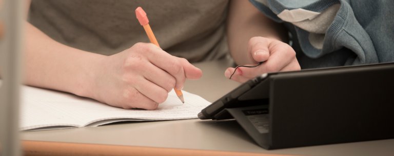 Hånd som holder blyant og skriver, en iPad i forgrunnen. Noen studerer.