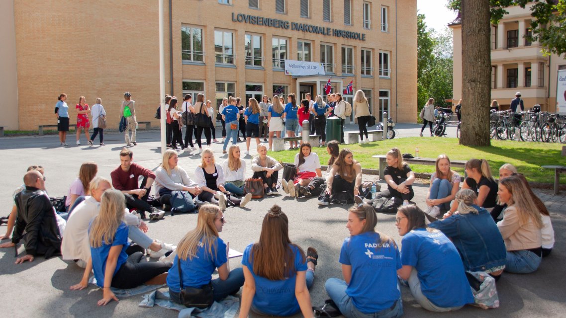 Studenter sitter i ring med faddere ute på plassen foran høyskolen