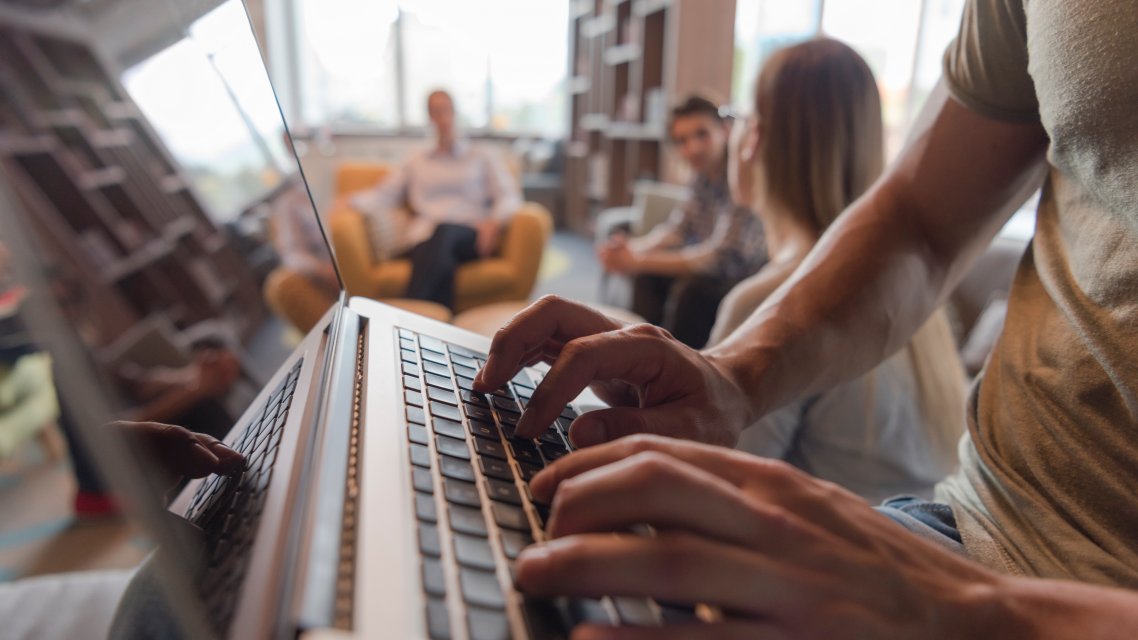 Hender som skriver på laptop, med en gruppe som sitter rundt